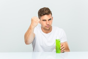 Young handsome man holding a aloe vera bottle showing fist to camera, aggressive facial expression.