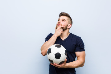 Young caucasian man holding a soccer ball looking sideways with doubtful and skeptical expression.