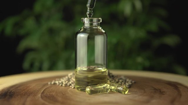Filling up glass jar bottle with cannabis cbd oil mixture for oral medical use. Made of green marijuana hemp plant on the background. Various organic medicine on the wood circle tree ring table.