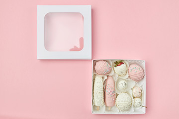 Ripe fruits covered with pink and white chocolate lie in a box on a pink background. View from above