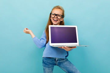 Copyspace photo of cheerful mischievous schoolgirl holds a notebook in her hands and gestures to the screen, a half-length shot against a blue wall