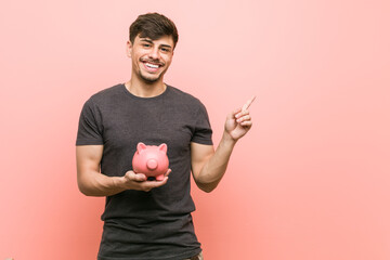 Young hispanic man holding piggy bank smiling cheerfully pointing with forefinger away.