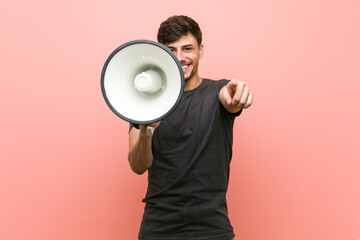 Young hispanic man holding a megaphone cheerful smiles pointing to front.