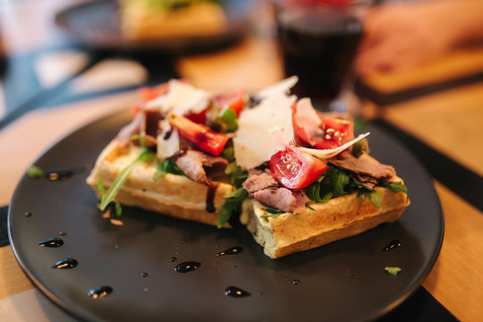 Waffles sandwich with bacon chiken and frech salad on black plate. Background of wooden table