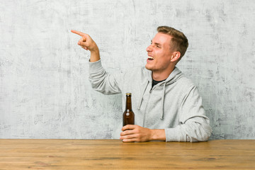 Young man drinking a beer on a table excited pointing with forefingers away.