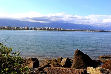 Rocks in the foreground, the  calm blue sea and a city in horizon. Blue sky and clouds. Bertioga, Brazil