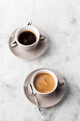 Two white cups of hot black coffee with milk isolated on bright marble background. Overhead view,...