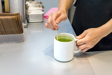 Female barista hand stirring matcha green tea on a mug with a stick at a coffe-shop conter in Hiroshima.