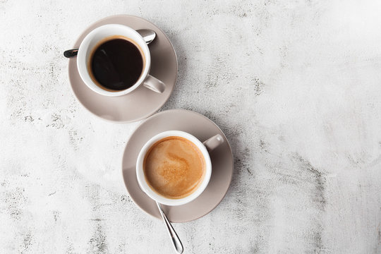 Two White Cups Of Hot Black Coffee With Milk Isolated On Bright Marble Background. Overhead View, Copy Space. Advertising For Cafe Menu. Coffee Shop Menu. Horizontal Photo.