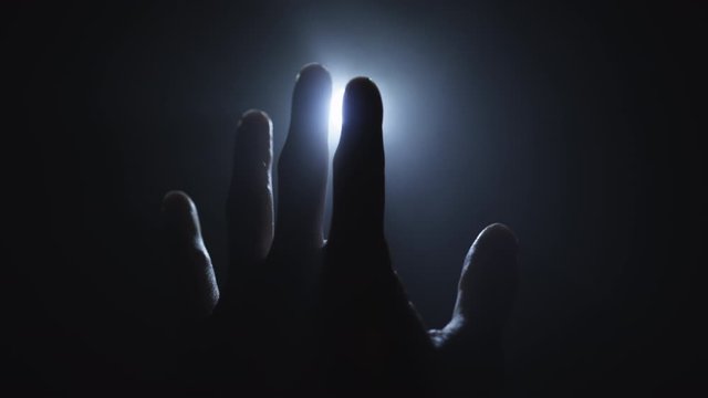 A mans hand reaches towards the light and fingers play with the light and pass over the source. Religion, spirituality and magic concepts.