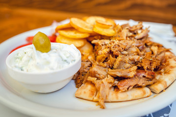 Close up view on gyros in white plate with pork meat pig and potato chips and tzatziki salad and tomato on the table