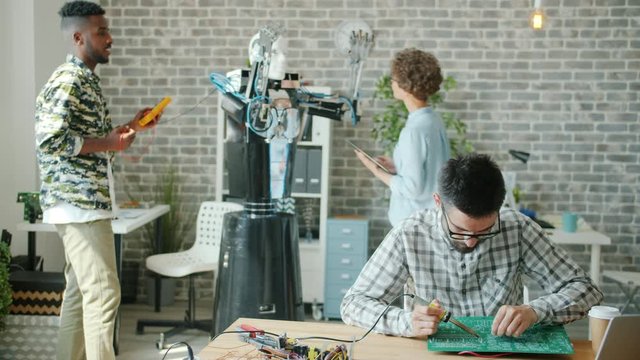 Multi-ethnic team of engineers is working in office with robot testing droid soldering circuit board with solderer. Robotics, engineering and modern technology concept.