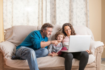 Child, a woman and a man sitting on a sofa watching a laptop and a smart phone.