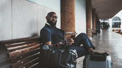 A mature African bald bearded man entrepreneur waiting for a train to start his business trip and sitting on the wooden bench at the platform of a railway station with a smartphone and bags near him