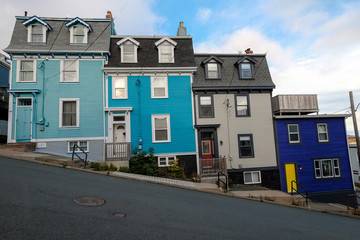 Multi-colored row houses on a street with a steep incline. The colorful houses have three levels. They have hip roofs and front doors on the exterior of the homes. The sky is blue with some clouds. 