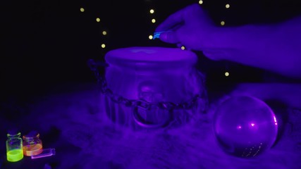 Halloween Bubbling smoking witch's cauldron with neon glowing vials and crystal ball in foreground. A hand empties poison into the pot. Witch and wizard concepts. RED Camera, Slow motion. - Powered by Adobe