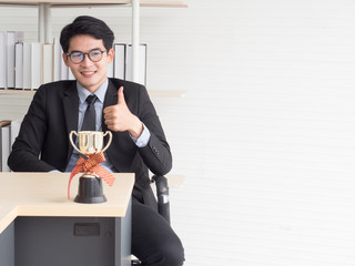 At the company, the office of an employee, a young businessman sat on a chair behind a steep, placed a book with a trophy placed in front of a golden color showing the achievements honored by the team