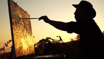 Professional artist paints a picture in nature. Silhouette of an adult man, sunset, abstract autumn landscape. Painting in the open air using a portable easel and a paintbox