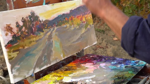 Oil painting on canvas. Professional artist adult man paints a picture in nature. Painting in the open air using a portable easel and a paintbox