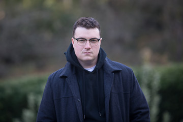 A caucasian male wearing a black hoodie sweater and glasses poses in a garden while surrounded by fall foliage on a cold afternoon.