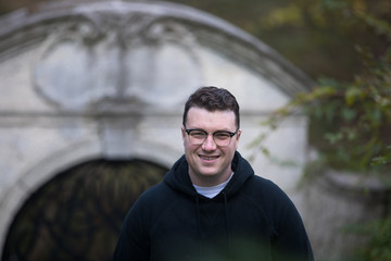 A caucasian male wearing a black hoodie sweater and glasses poses in a garden while surrounded by fall foliage on a cold afternoon.