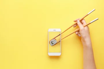 Foto op geborsteld aluminium Sushi bar Vrouwelijke hand met eetstokjes en mobiele telefoon met smakelijke sushi roll op het scherm tegen een achtergrond met kleur