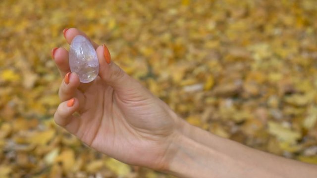 Female hand with orange manicure holding transparent violet amethyst yoni egg for vumfit, imbuilding or meditation on yellow fallen leaves background during autumn day outdoors