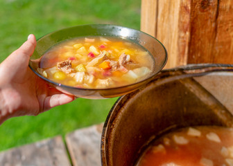The girl's hands outdoors holding a transparent plate of soup borscht with meat and vegetables on the background of a cauldron with food in nature in a wooden house.