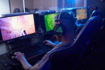 Fototapeta na wymiar Rear view of young gamer wearing gaming headphones with backlight and playing in computer video game on computer in dark computer class
