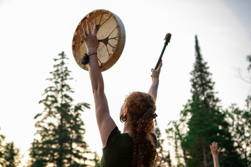 A spiritual redhead women is viewed from behind as she raises a native drum and drumstick with...
