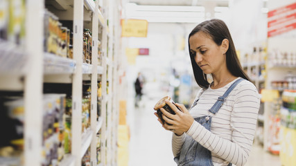 Young woman in denim overall is selecting canned mushrooms in grocery store. Customer chooses goods...