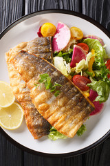 fried sea bass fillet with lemon and fresh vegetable salad close-up on a plate. Vertical top view