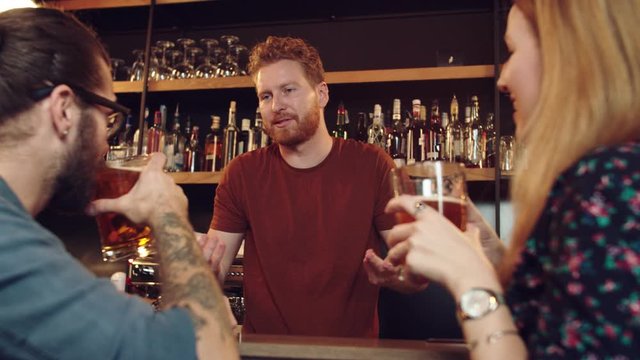 Cheerful friends leaning on bar counter, drinking beer and chatting with bartender. Night out.