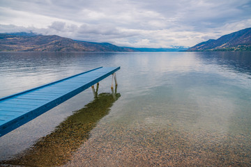 Blue painted dock extending over calm lake with view of overcast sky and mountains