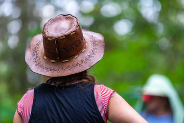 Selective focus close up of woman wearing cowboy leather hat viewed from the back, with blurred background of nature and people