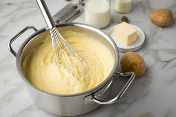 Kochtopf mit Kartoffelpüree mit Zutaten aus Kartoffeln, Milch, Salz, Butter, Muskatnuss dazu Muskatreibe und Schneebesen auf hell Marmor Hintergrund