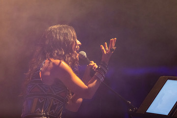 a female musician is viewed from the side as she sings, with open mouth in microphone, with smoke...