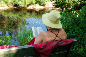 old woman is seen from behind as she reading a book in beautiful nature, wearing a summer hat and enjoying a beautiful sunny day by the river