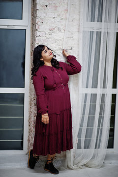 Attractive south asian woman in deep red gown dress posed at studio against windows in night.