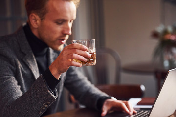 Elegant young guy in formal wear sits in cafe with his laptop and glass of alcohol