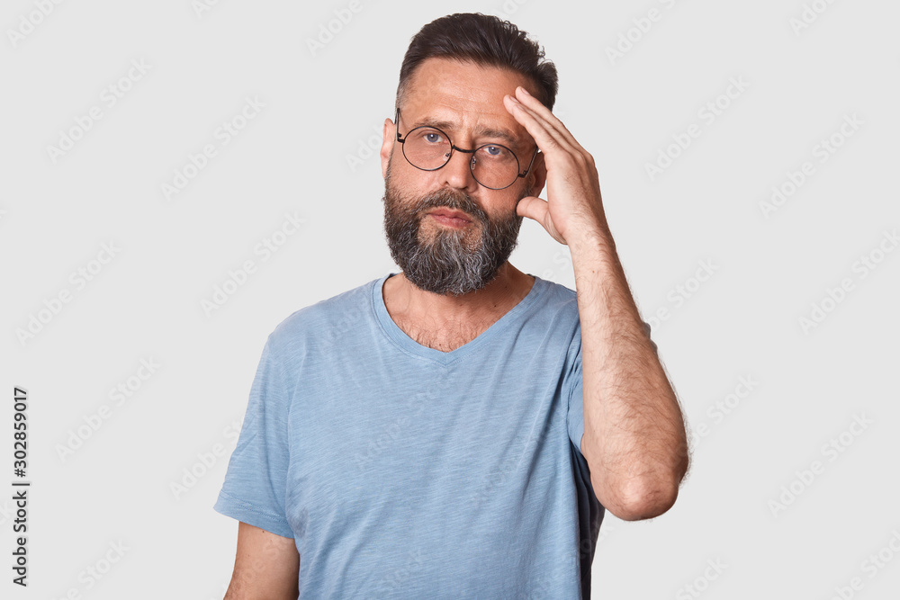 Wall mural portrait of serious handsome male with beard and mustache, keeps hand on forhead, wearing grey t shi