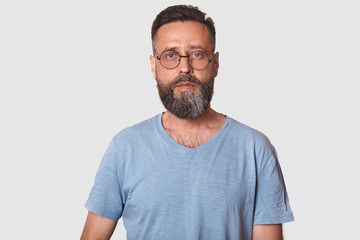Close up portrait of middle aged male wearing eyeglasses and caual gray t shirt, looking directly at camera, posing against white background with copy space, attractive guy with beard. People concept.