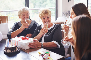 Glasses with alcohol in hands and cake on table. Senior woman with family and friends celebrating a birthday indoors