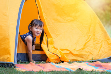 Little Asian girl smile and looking at camera playing with her tent on campsite, happy girl sitting inside yellow tent at park