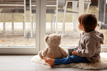 Cute little toddler boy sitting at window with cup of warm tea with his soft teddy bear toy looking...