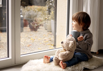 Cute little toddler boy sitting at window with cup of warm tea with his soft teddy bear toy looking out thoughtfully at chilly autumn weather. Cozy home. Fall melancholy concept. Seasonal mood
