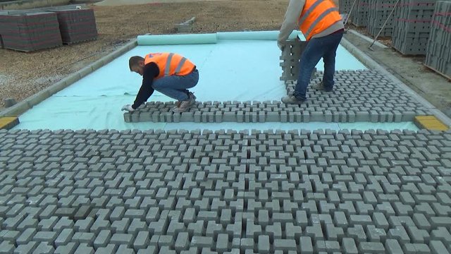 Construction worker laying interlocking paving concrete onto sheet nonwoven bedding sand and fitting them into place.