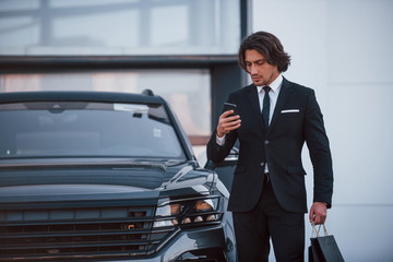 Portrait of handsome young businessman in black suit and tie outdoors near modern car and with shopping bags
