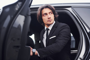 Fototapeta na wymiar Portrait of handsome young businessman in black suit and tie walks out from modern car