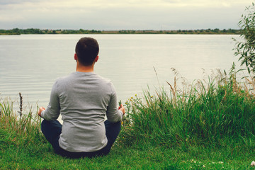 Fototapeta premium A young guy in a light polo sits on the shore. The view from the back. Yoga training. Appeasement.